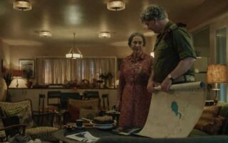 Vintage film set with two actors discussing a map in a cosy living room.