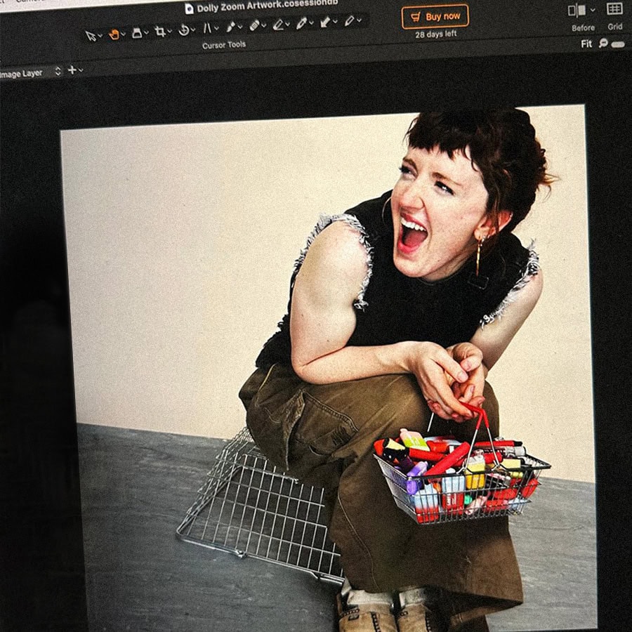 A woman with short dark hair laughing while sitting on a metal shopping trolley with a basket of col.