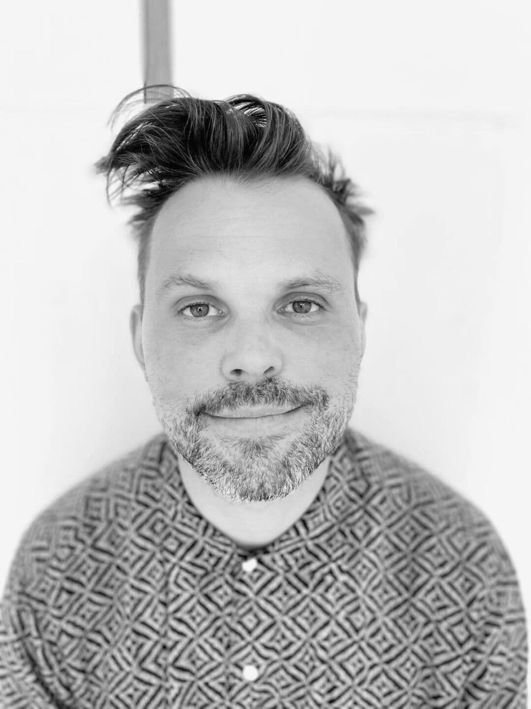 PHOTO-2026-04-28-14-24-27 A man with styled hair and a beard, wearing a patterned shirt, smiling at the camera in black and wh.