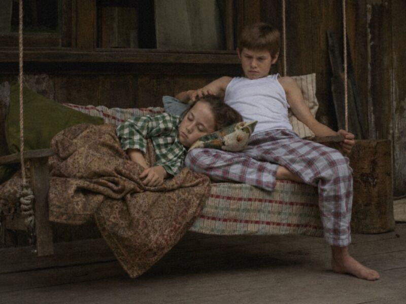 Two children resting on a rustic wooden bed in a cozy cabin setting.